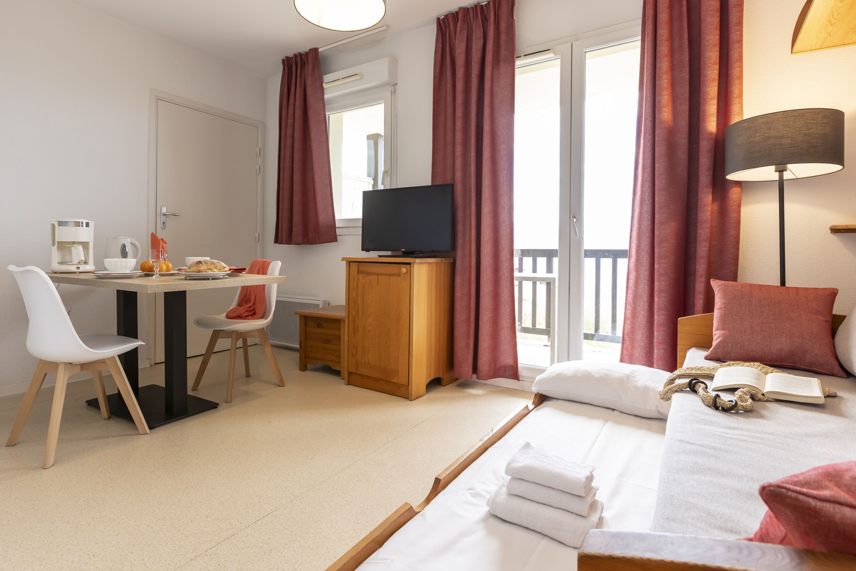 Une chambre d’hôtel comprenant un lit, un bureau avec une télévision, une chaise, une machine à café et une fenêtre avec des rideaux.
