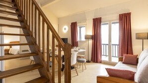 A hotel room with a wooden staircase, a dining area with a table and chairs, a sofa with cushions, and a large window with curtains. - Résidence Odalys Mille Soleils (Font-Romeu-Odeillo-Via)