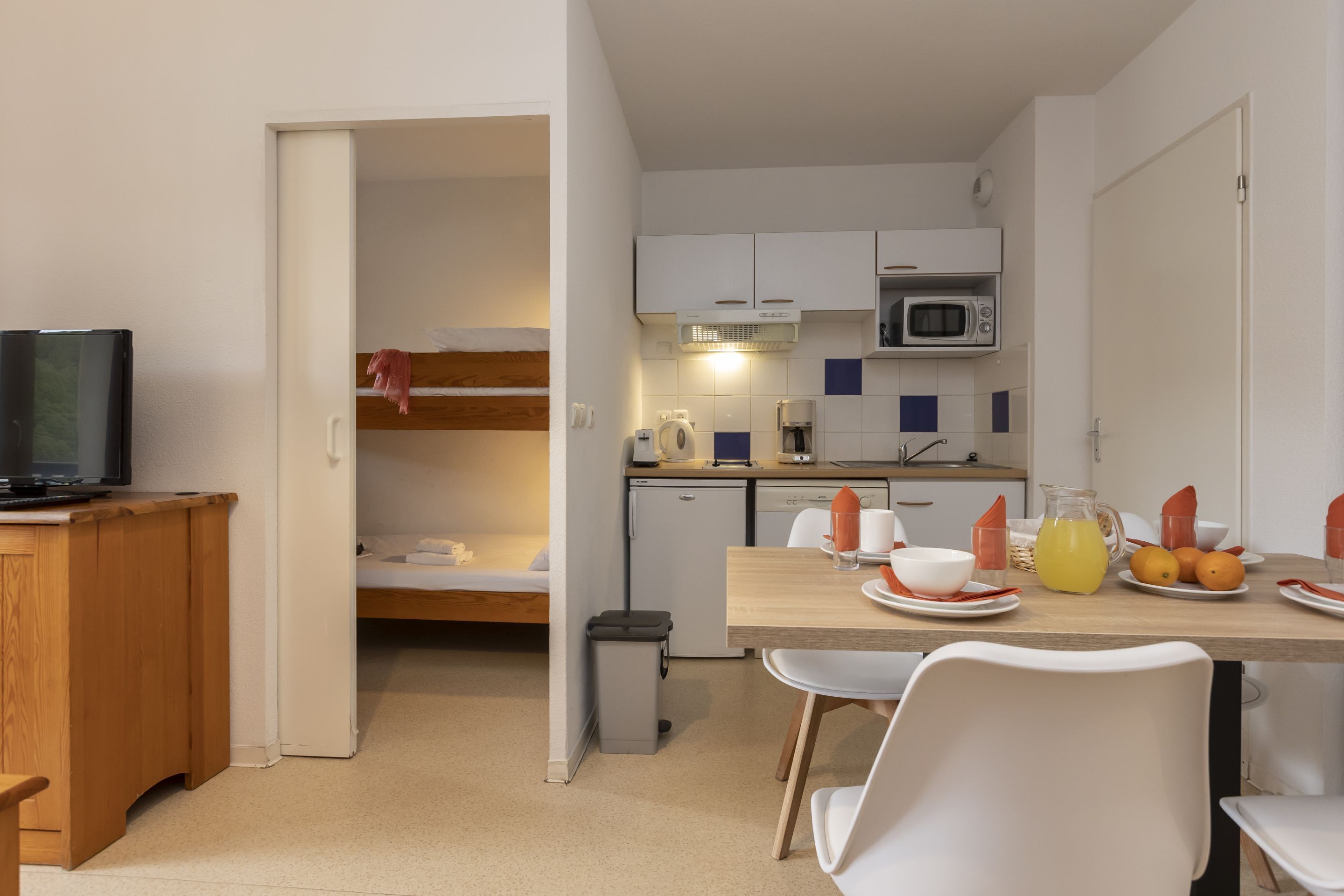 A compact kitchen with a microwave, coffee maker, and a small refrigerator. There is a dining table with chairs and a TV on a wooden cabinet.