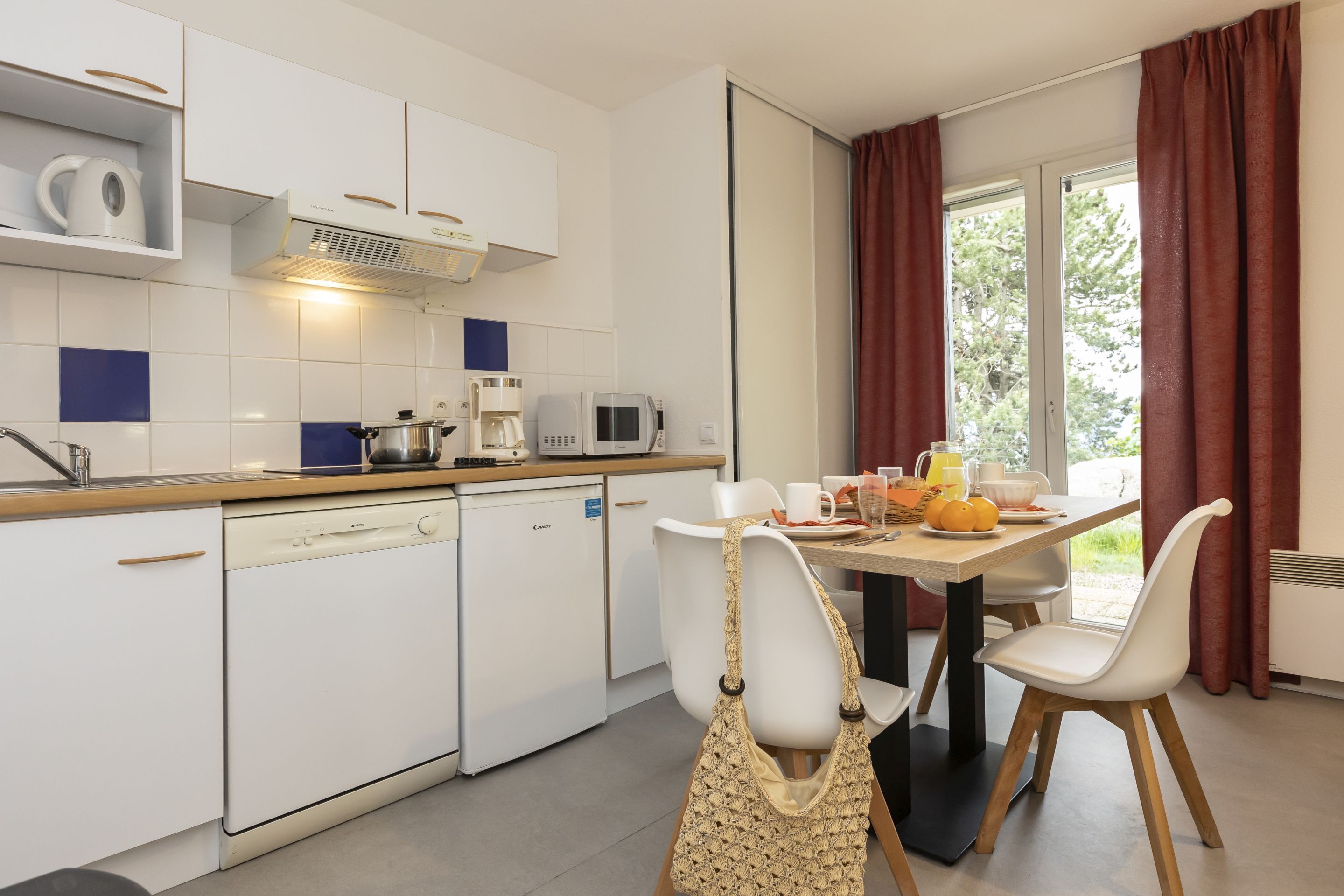 Cocina compacta con armarios blancos, lavavajillas, microondas y cafetera. Hay una mesa de comedor con sillas y una ventana con cortinas.