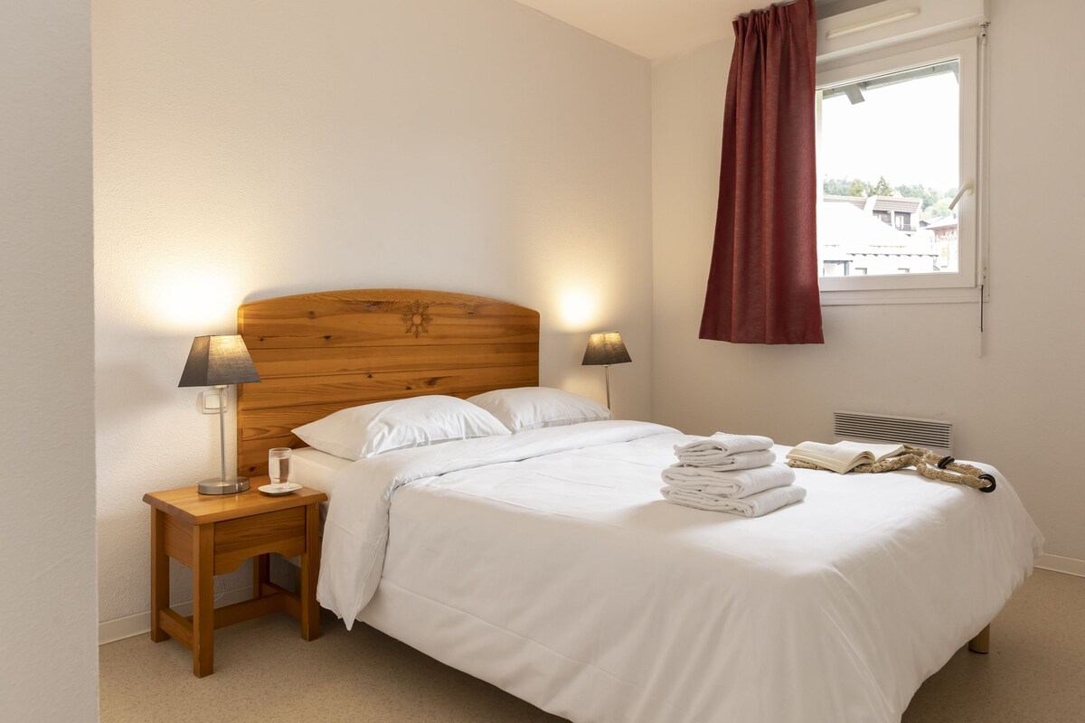 A neatly made bed with white linens, two bedside lamps, a window with a view of buildings, and a stack of folded towels on the bed.