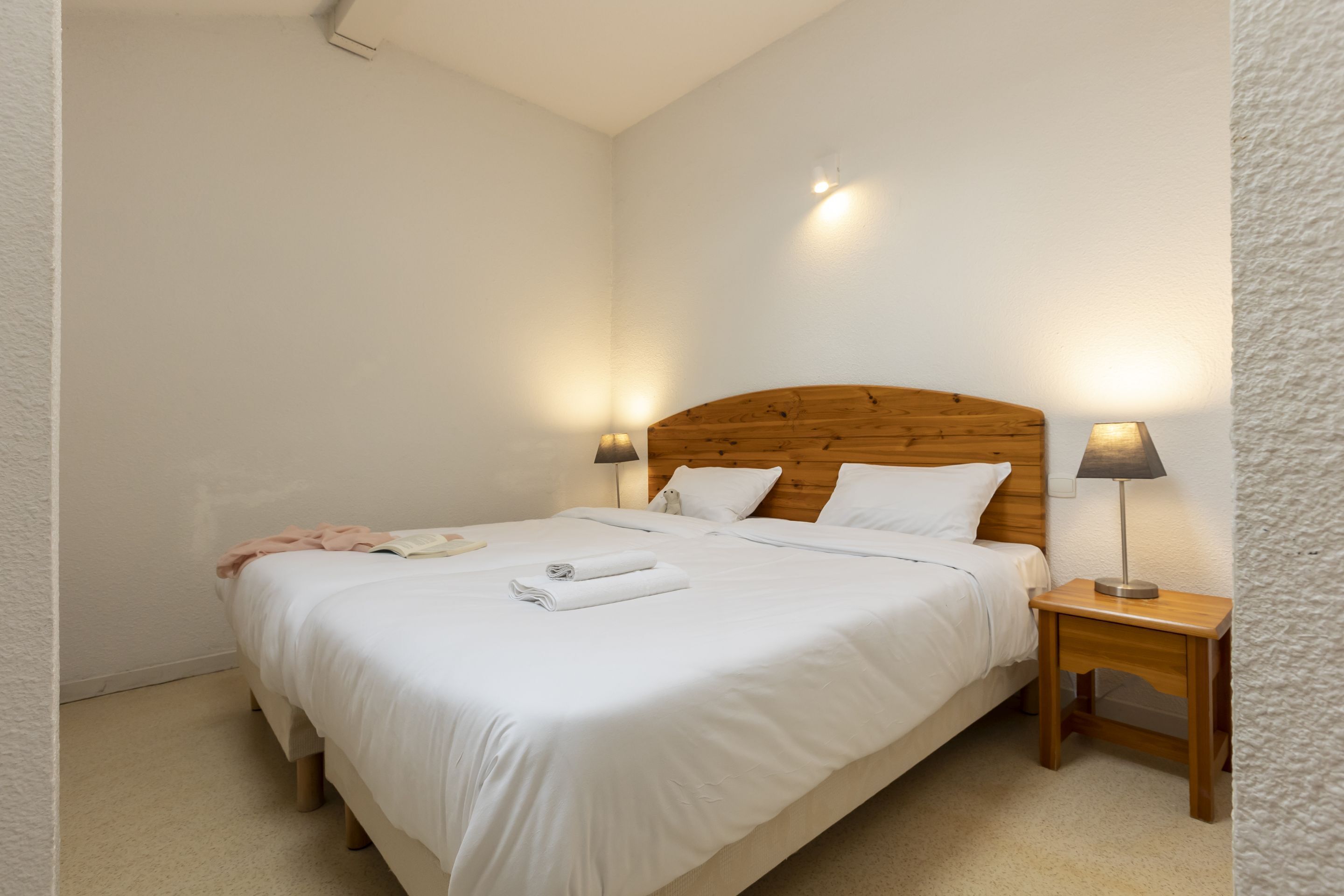 A neatly made bed with white linens and a folded towel, a wooden headboard, two bedside lamps, and a small wooden nightstand.