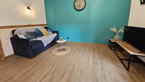 A living room with a teal wall, a dark blue sofa, a small round table, a TV on a wooden stand, and a decorative clock. - Gite "Bleuet", the little LOFT, 2 to 6 persons with SPA in the Residence  (Ventron)