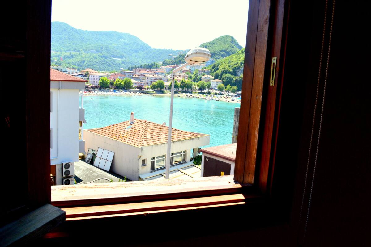 A view from a window overlooking a coastal town with buildings, a beach, and mountains in the background.