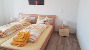 Ein Doppelbett mit gemustertem Überwurf und Kissen, ein Nachttisch mit Lampe sowie ein Wandbild mit Sonnenuntergang über Wasser.