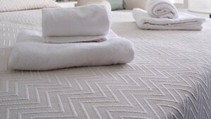 Een net opgemaakt bed met opgevouwen handdoeken, een stoel met een kussen en een lijst met een afbeelding aan de muur.
