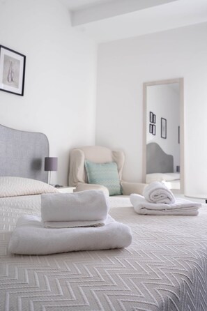 Ein ordentlich bezogenes Bett, gefaltete Handtücher, ein Sessel mit Kissen und ein gerahmtes Bild an der Wand.