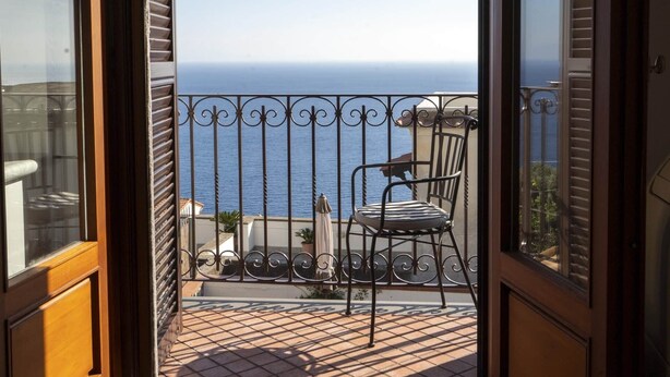 Een open schuifdeur die leidt naar een balkon met uitzicht op zee, een stoel en een klein tafeltje.