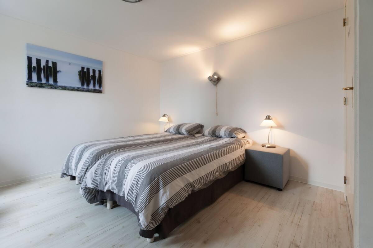 A bedroom with a bed, two bedside tables with lamps, a wall-mounted light, and a painting on the wall.