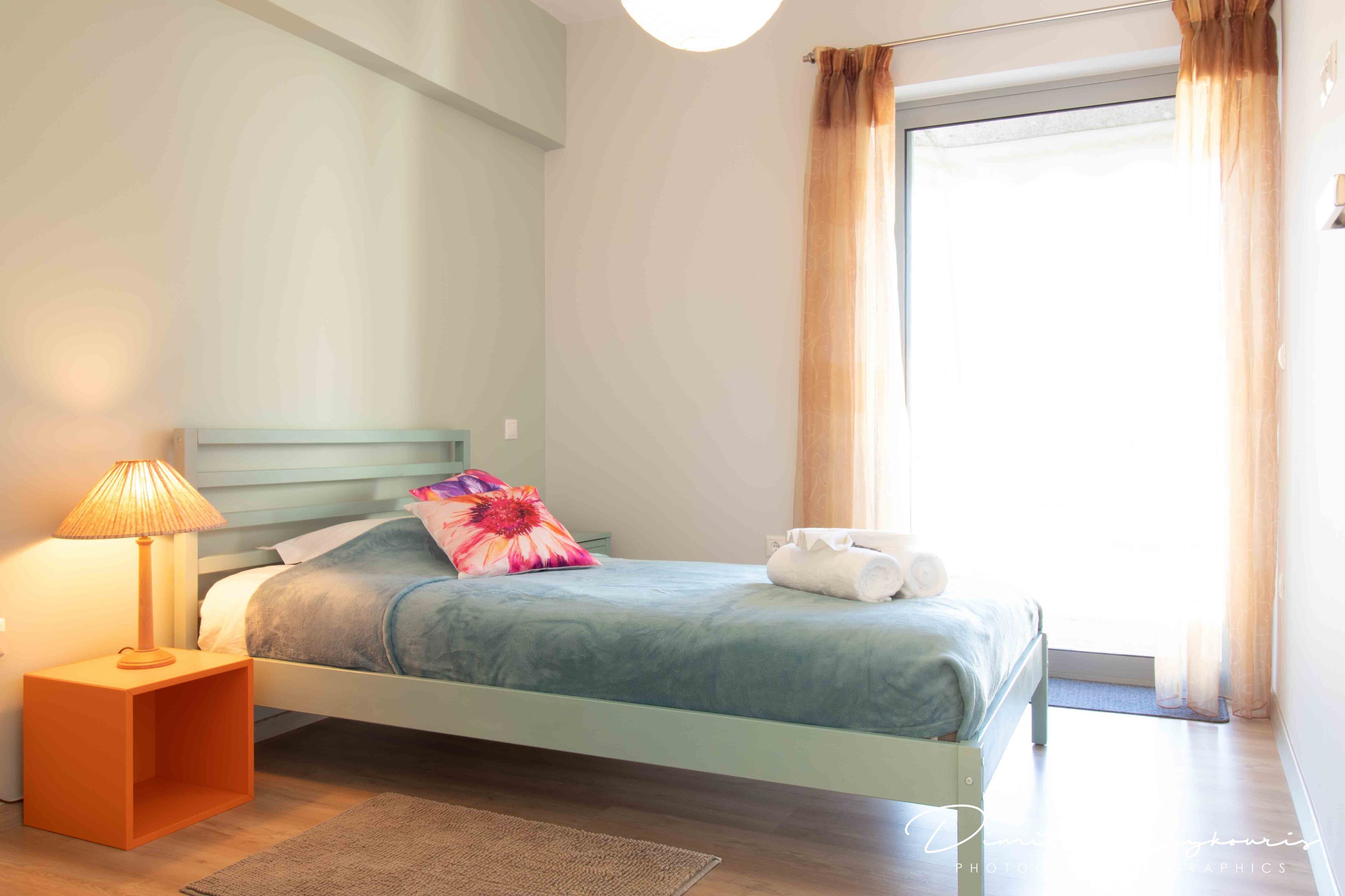 Ein ordentlich bezogenes Bett mit blauer Bettdecke und farbigem Kopfkissen, eine Nachttischlampe und ein kleiner Nachttisch.