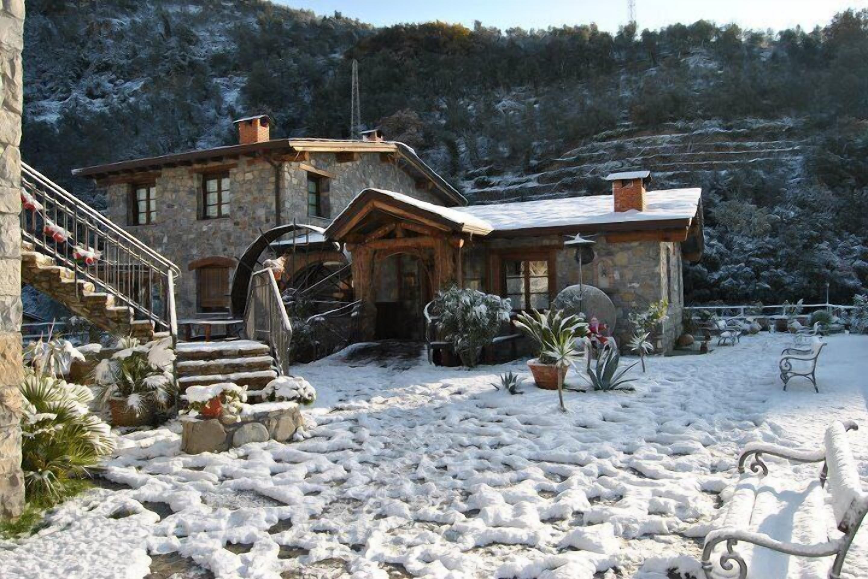 A stone house with a wooden staircase leading to the entrance, surrounded by snow-covered ground and plants.