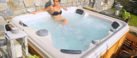 A hot tub with a stone wall backdrop, a small table with a lantern, and a wooden deck.