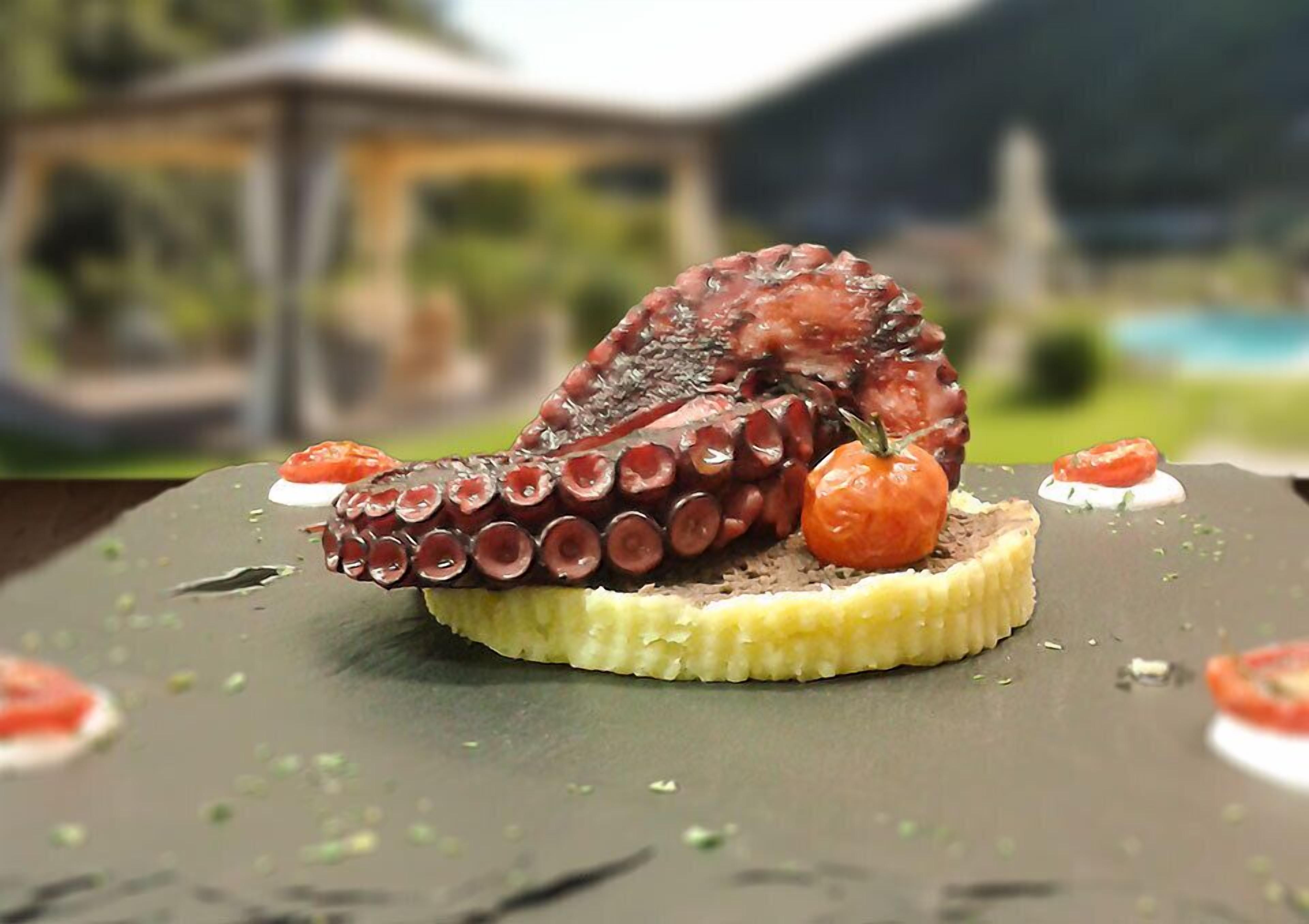 Un plato de postre con una rebanada de pastel coronada con un tentáculo de pulpo y tomates cherry.