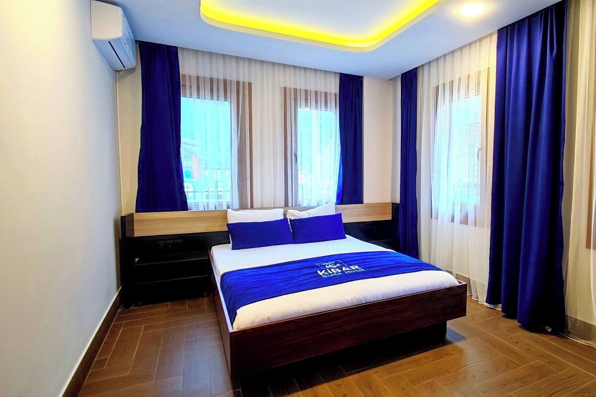 A modern hotel room with a large bed, wooden headboard, and a view of the outdoors through sheer curtains.