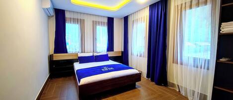 Quarto de hotel moderno com uma cama grande, cabeceira de madeira e vista para o exterior através de cortinas leves.