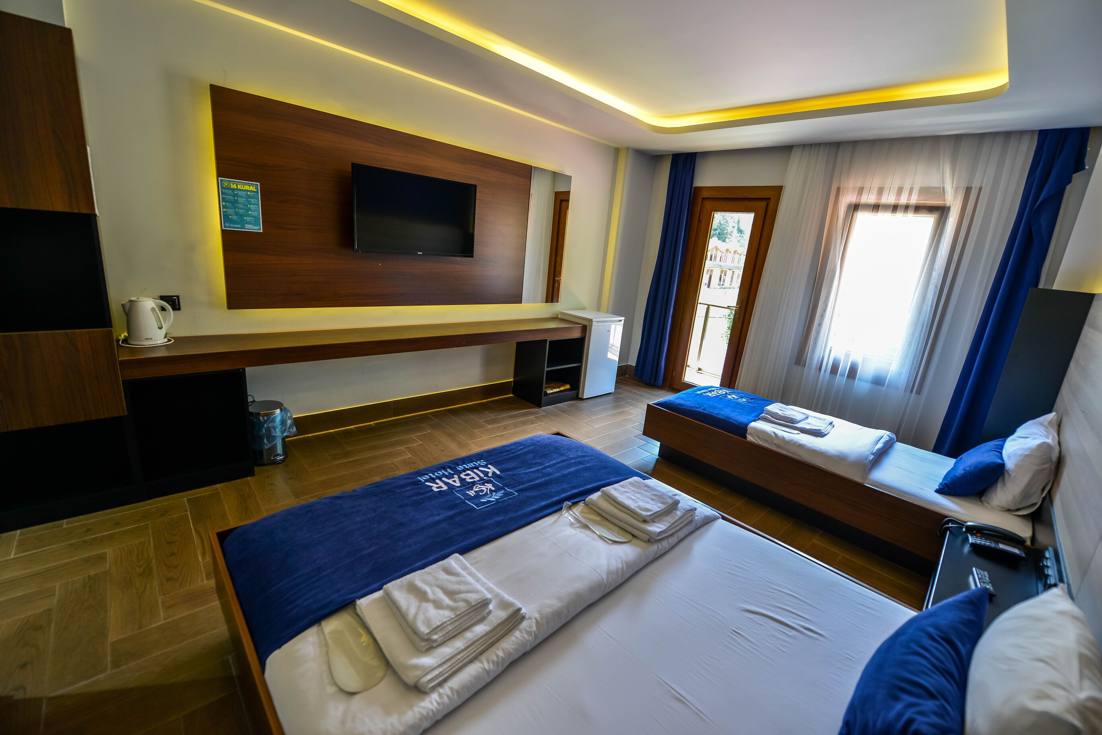Habitación de hotel con cama, sofá cama, televisor montado en la pared, mesita con tetera y ventana con cortinas.