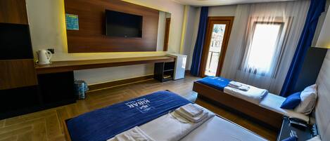 Habitación de hotel con cama, sofá cama, televisor montado en la pared, mesita con tetera y ventana con cortinas.
