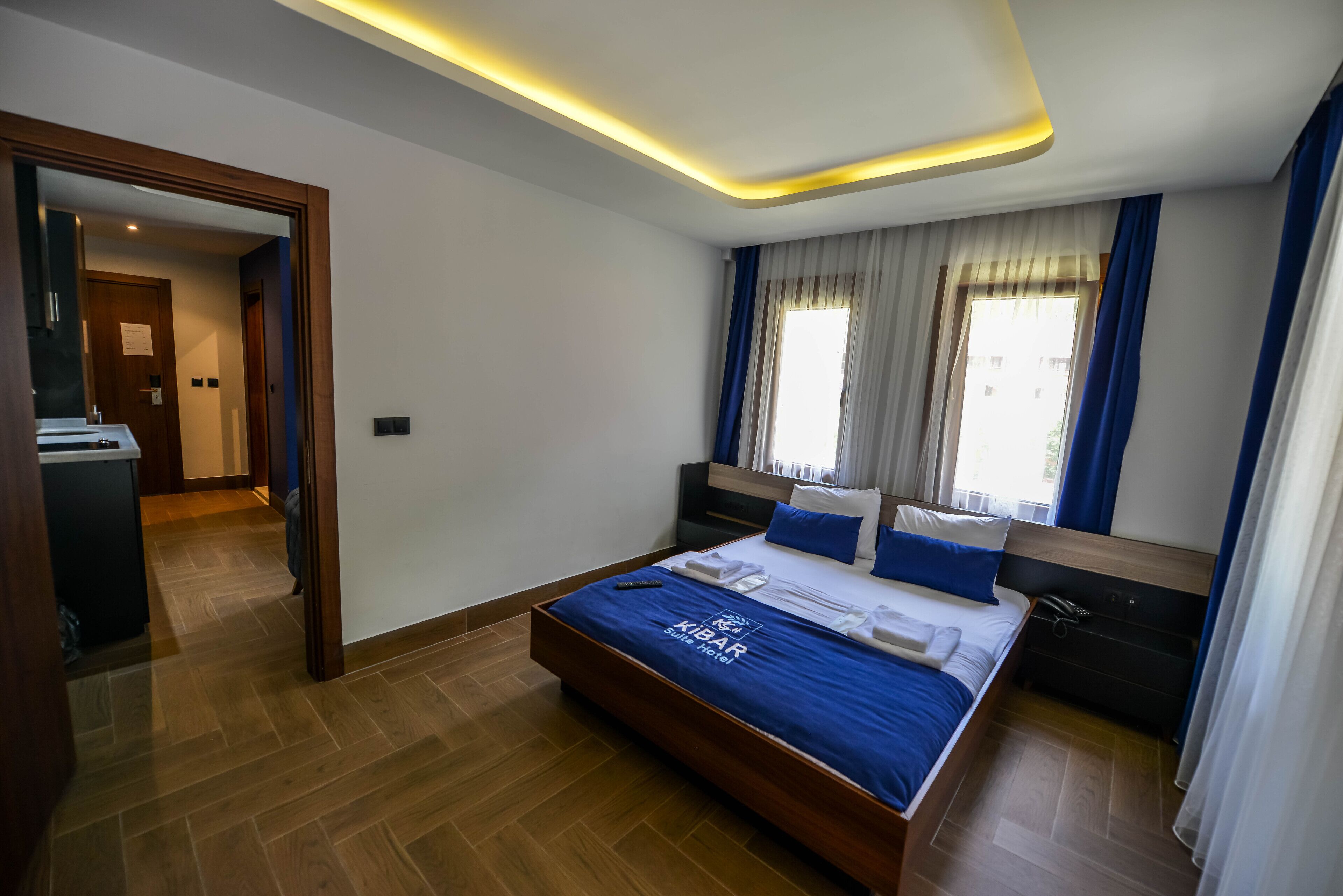 Habitación de hotel con cama, mesita de noche, espejo, ventana con cortinas y suelo de madera.