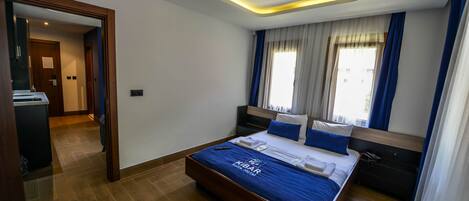 Habitación de hotel con cama, mesita de noche, espejo, ventana con cortinas y suelo de madera.