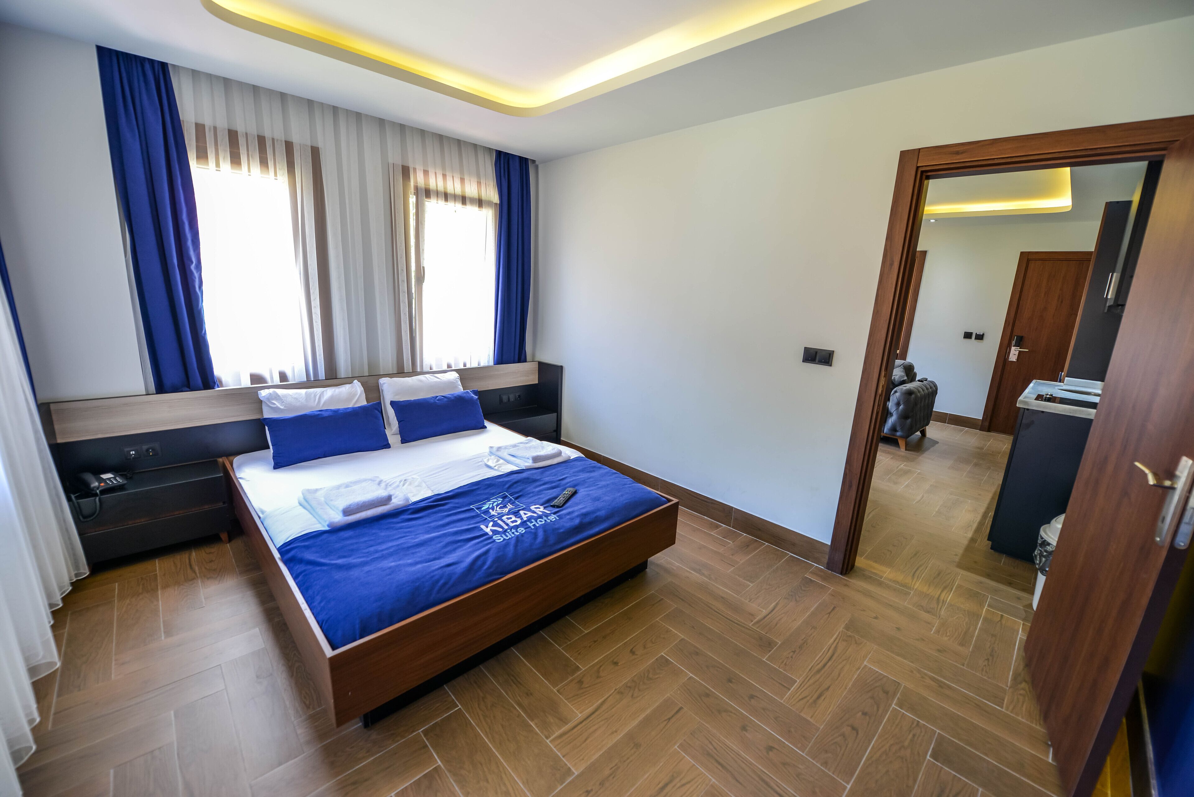 A modern hotel room with a large bed, wooden headboard, and a blue bedspread. There is a wooden floor, a small desk with a chair, and a mirror on the wall.