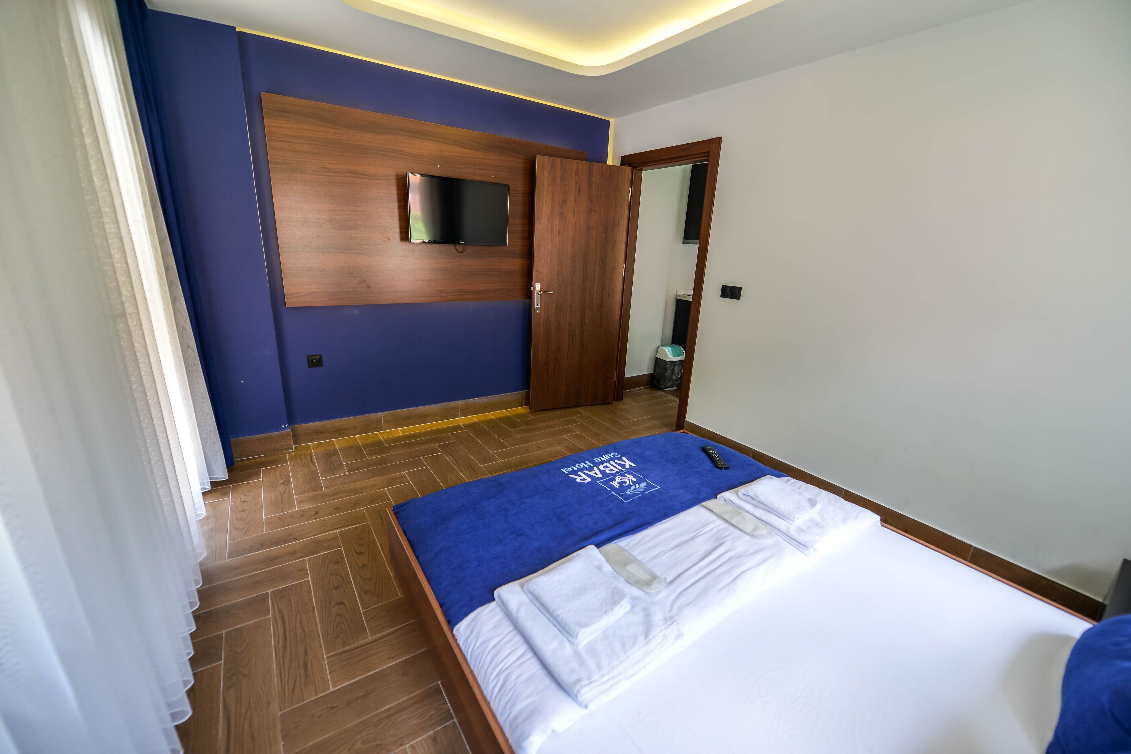A hotel room with a wooden floor, a bed with a blue bedspread, and a TV mounted on the wall.