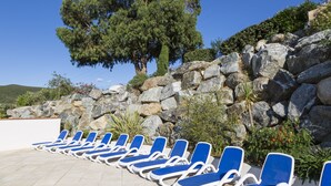 A row of blue lounge chairs with white frames, arranged on a tiled patio area with a rocky backdrop and a large tree. - Odalys Residence Les Hameaux de Capra Scorsa (Belgodere)