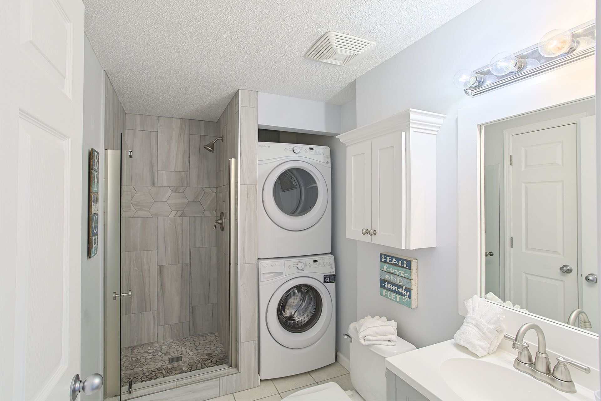 Une petite buanderie comprenant deux machines à laver superposées, un espace douche et une salle de bain avec un lavabo et un miroir.