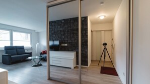 A modern living room with a glass partition, a sofa, a coffee table, a TV, and a tripod in the hallway. - Business Apartments Ratingen West (Ratingen)