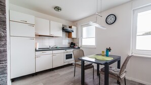 A modern kitchen with white cabinets, a dining table with chairs, a clock on the wall, and a window providing natural light. - Business Apartments Ratingen West (Ratingen)