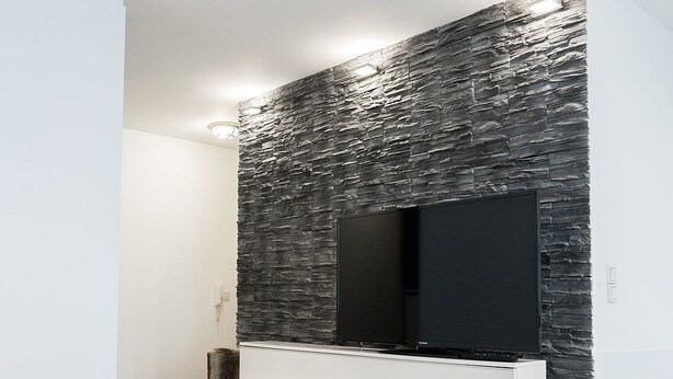 A modern living room with a flat-screen TV mounted on a textured wall, a white entertainment unit below, and a wooden floor. - Business Apartments Ratingen West (Ratingen)