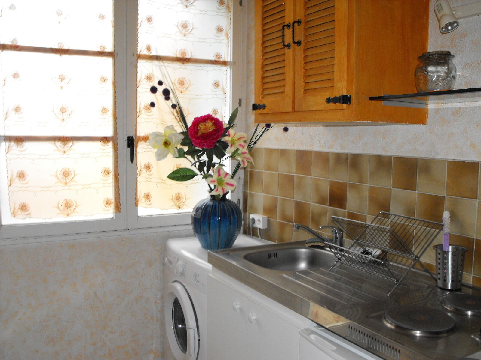 Une cuisine équipée d’un lave-linge, d’un évier muni d’une passoire et d’une fenêtre ornée d’un motif floral.