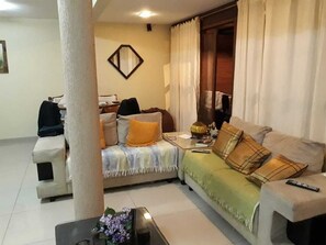 Ein Wohnzimmer mit einer Couch, einem Couchtisch und einem Sessel. Ein Fenster mit Vorhängen, ein Spiegel an der Wand und eine Tür im Hintergrund.