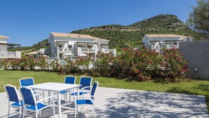 A patio with a white table and blue chairs, surrounded by a garden with pink flowers and a view of a hillside. - Résidence Odalys Casa d'Orinaju (Oletta)