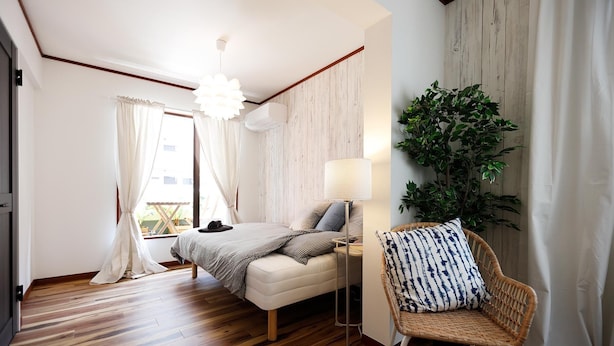 A bedroom with a bed, a bedside lamp, a wicker chair, and a large plant.