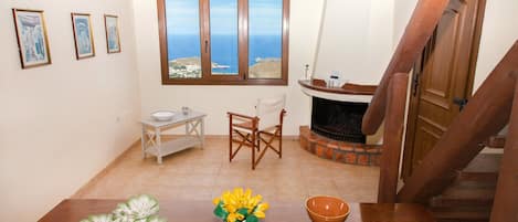 A dining area with a wooden table, a vase with yellow flowers, a bowl, and a window with a view of the sea.