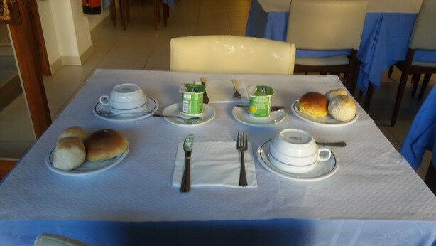 A dining table set with plates of bread rolls, a cup of tea, a green container, and a teapot. - Hotel Riomar (Lagos)