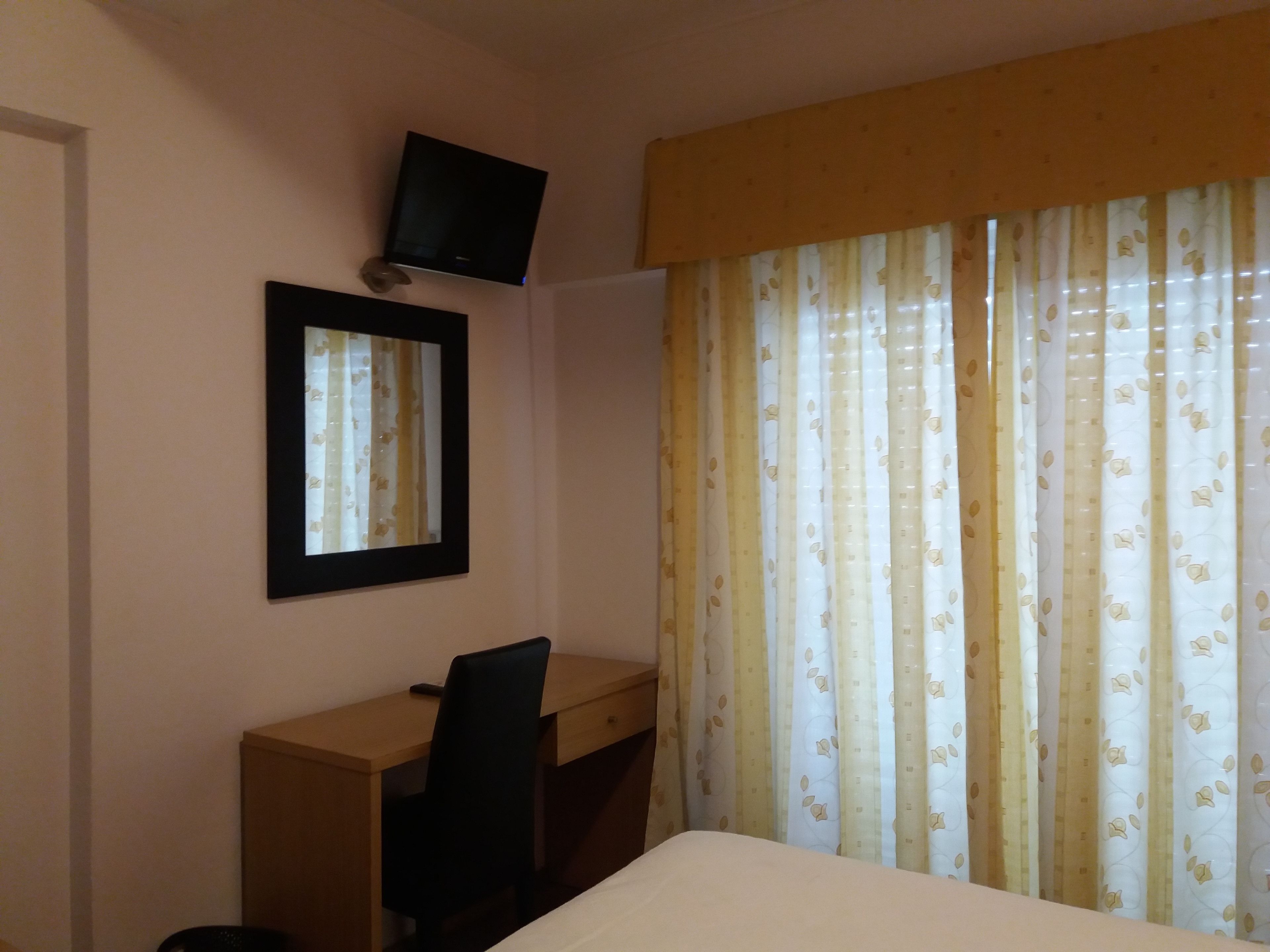 A hotel room with a bed, a desk, a chair, a television mounted on the wall, and a window with curtains.