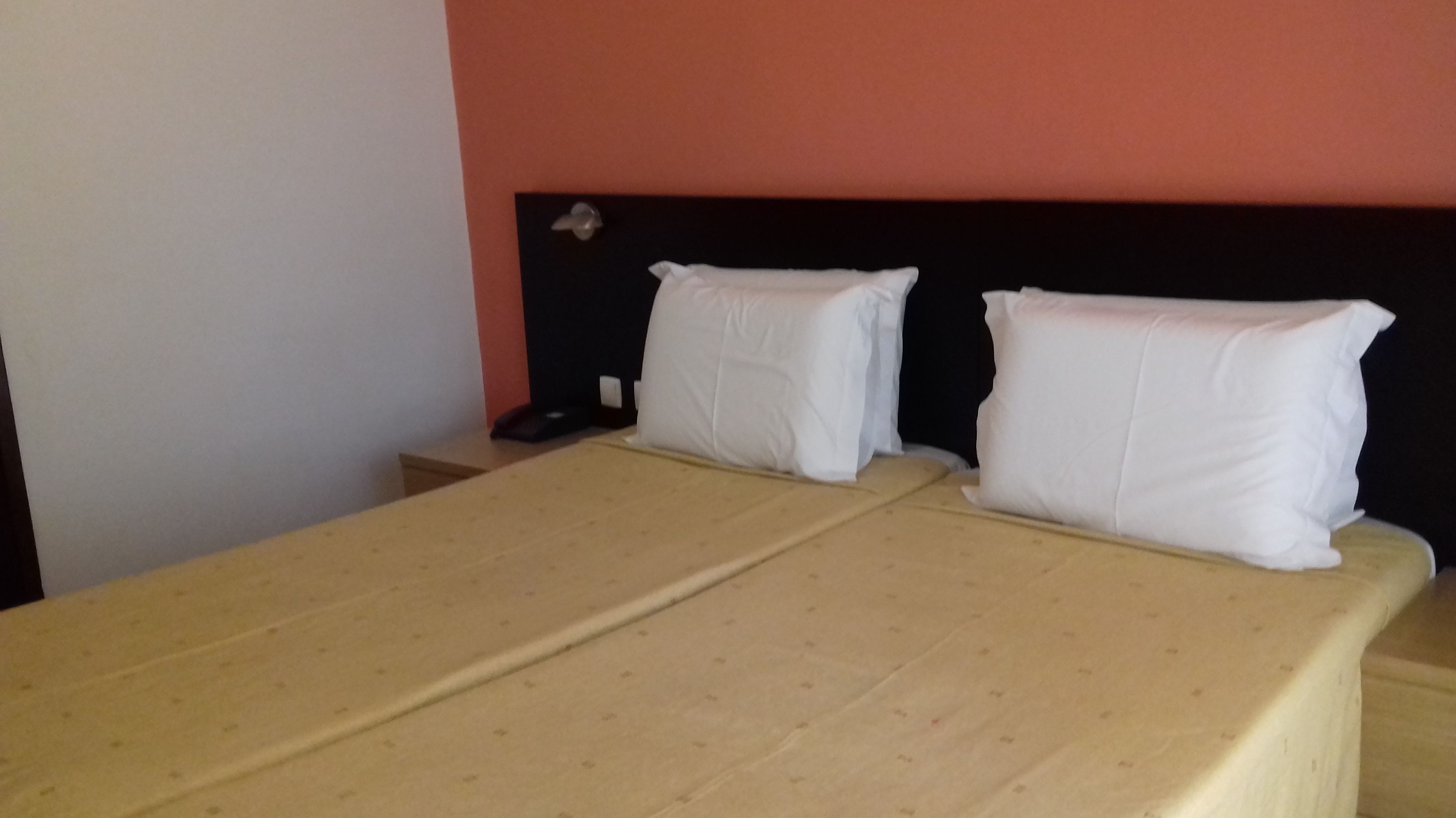 Una cama doble con una colcha color beige y dos almohadas blancas, una mesita de noche con un teléfono, y una pared con un cabecero rojo y negro.