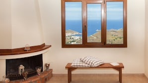 A room with a fireplace, a bench with cushions, and a window with a view of the sea and a distant town. - Villa Karthea IV (Kea)