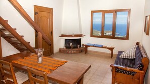 A cozy room with a wooden staircase, a fireplace, a dining table with chairs, and a sofa with a view of the sea. - Villa Karthea IV (Kea)