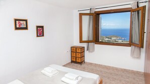 A bedroom with a bed, two framed artworks, a wooden chest, and a window with a view of the sea. - Villa Karthea IV (Kea)