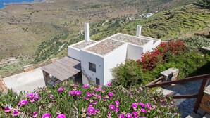 A white building with a flat roof, surrounded by a garden with pink flowers, overlooking a hilly landscape with a view of the sea. - Villa Karthea IV (Kea)