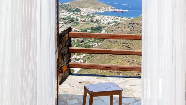 A balcony with a stone floor, a wooden chair, and a view of the sea and a distant town. - Villa Karthea IV (Kea)