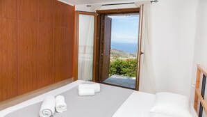 Ein Schlafzimmer mit einem Bett, zwei zusammengelegten Handtüchern, einem Holzschrank und einer Glastür, die auf einen Balkon mit Meerblick führt.