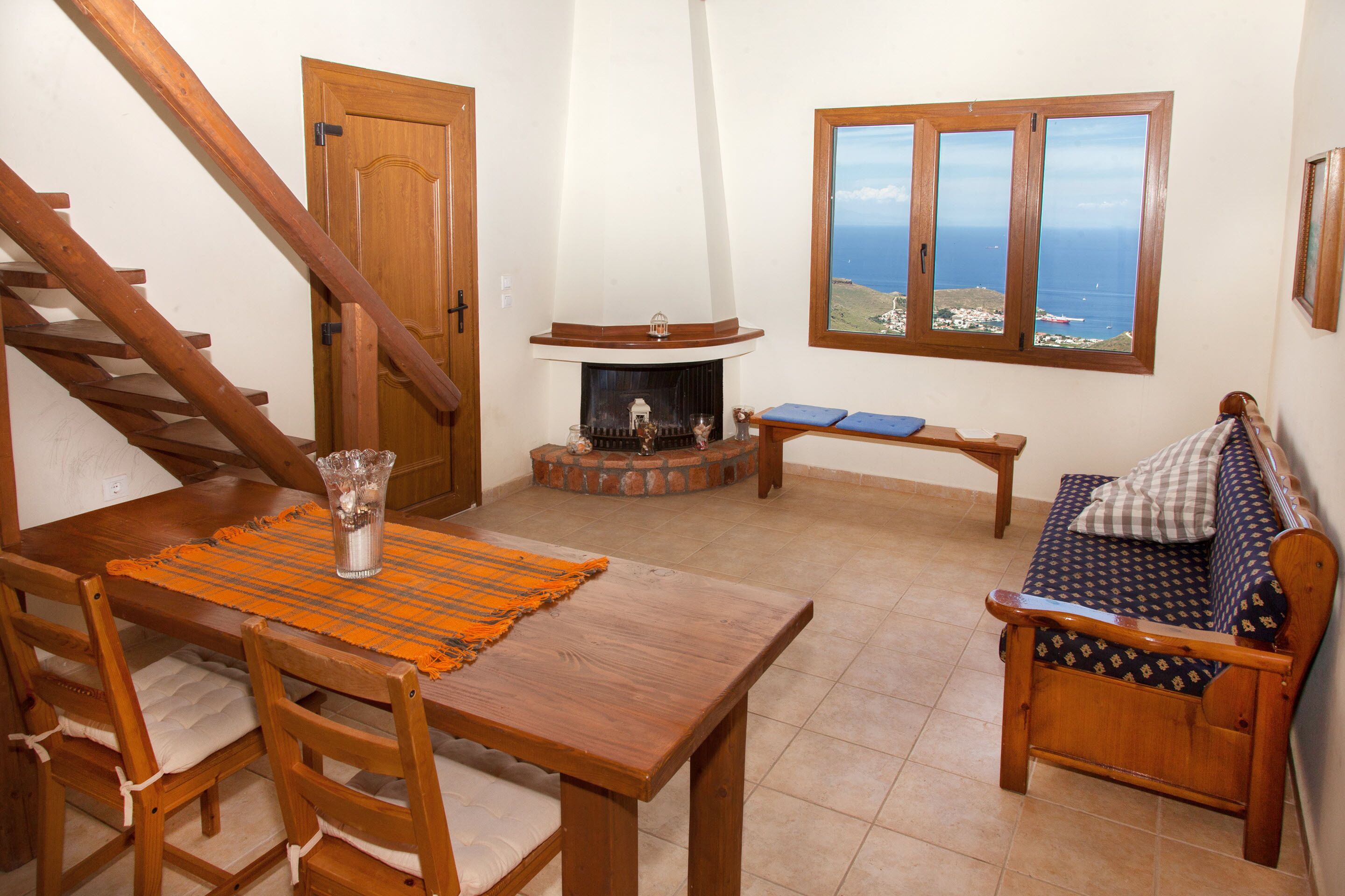 A cozy room with a wooden staircase, a fireplace, a dining table with chairs, and a sofa with a view of the sea.