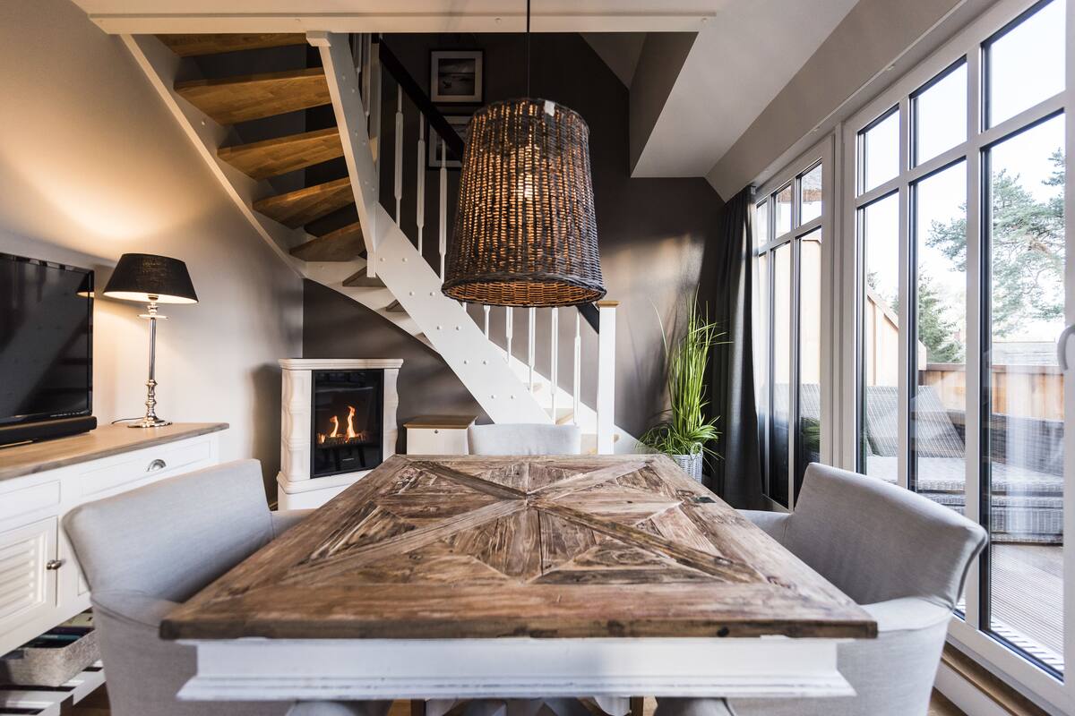 Un comedor con una gran mesa de madera, sillas blancas, una lámpara colgante de mimbre, una chimenea y una escalera con barandilla blanca.