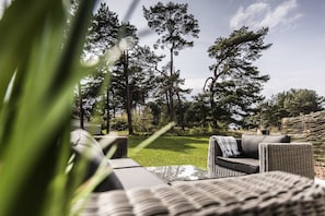 A patio area with wicker furniture and a glass table, surrounded by a grassy lawn and tall trees. - Kamphof Usedom Ferienanlage (Loddin)
