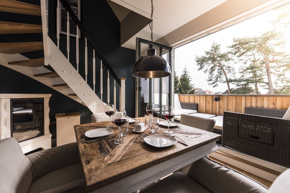 Un comedor con una mesa de madera dispuesta para dos personas, una escalera y vistas a árboles.