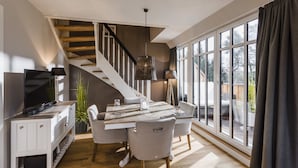 A modern living room with a wooden staircase, a dining area with a large window, and a TV on a white cabinet. - Kamphof Usedom Ferienanlage (Loddin)