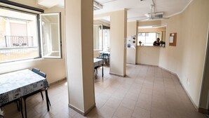 Une pièce spacieuse comprenant une table et des chaises, un balcon avec une rambarde, et un coin cuisine où l’on aperçoit une personne.